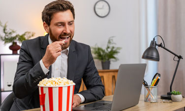 Middle-aged businessman at home office watches movie on laptop at lunch break and eats popcorn smiling. Freelancer happy guy at table laughs at funny scene eats popcorn and enjoys break before work.